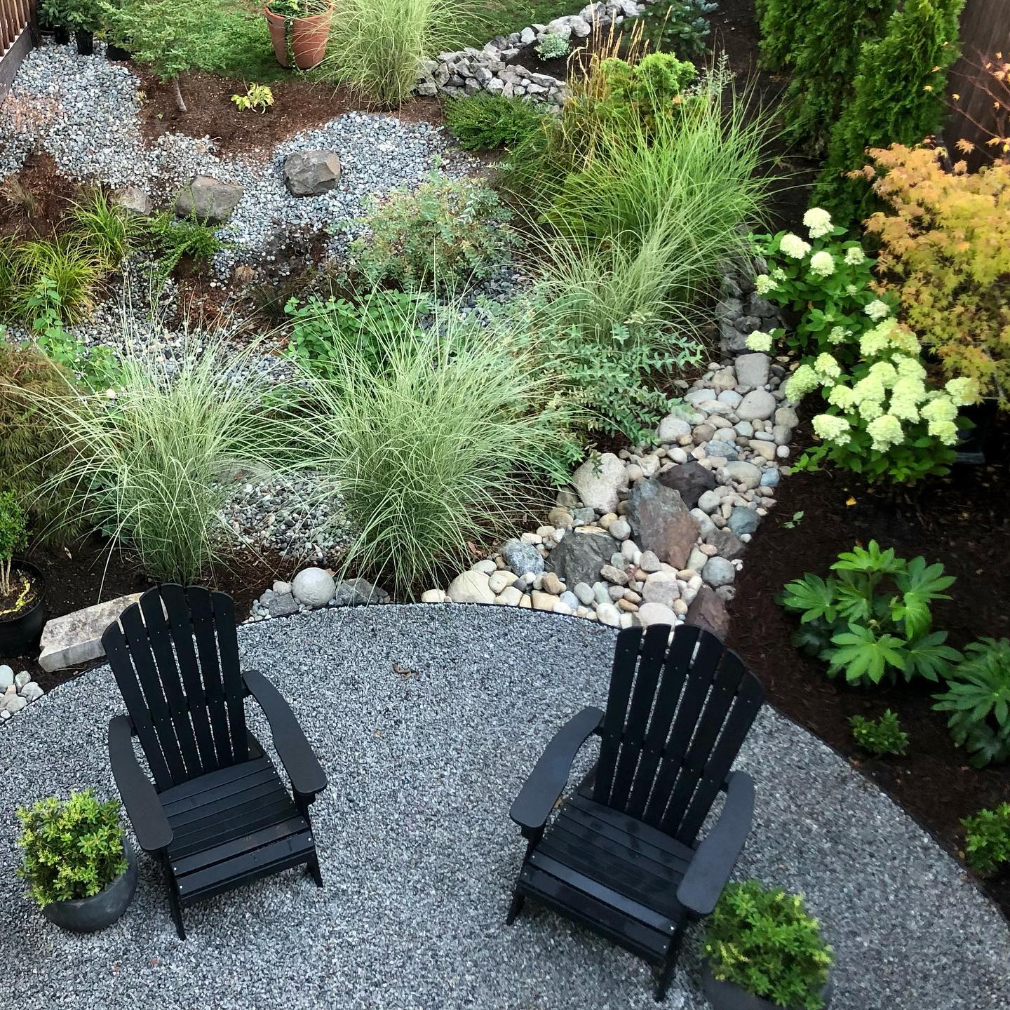 A patio with two black chair and grasses and flowers around.